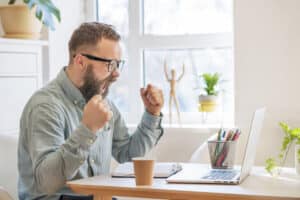 Happy businessman feels excitement, screams, and raises his fists while looking at his laptop in a celebration of his success.