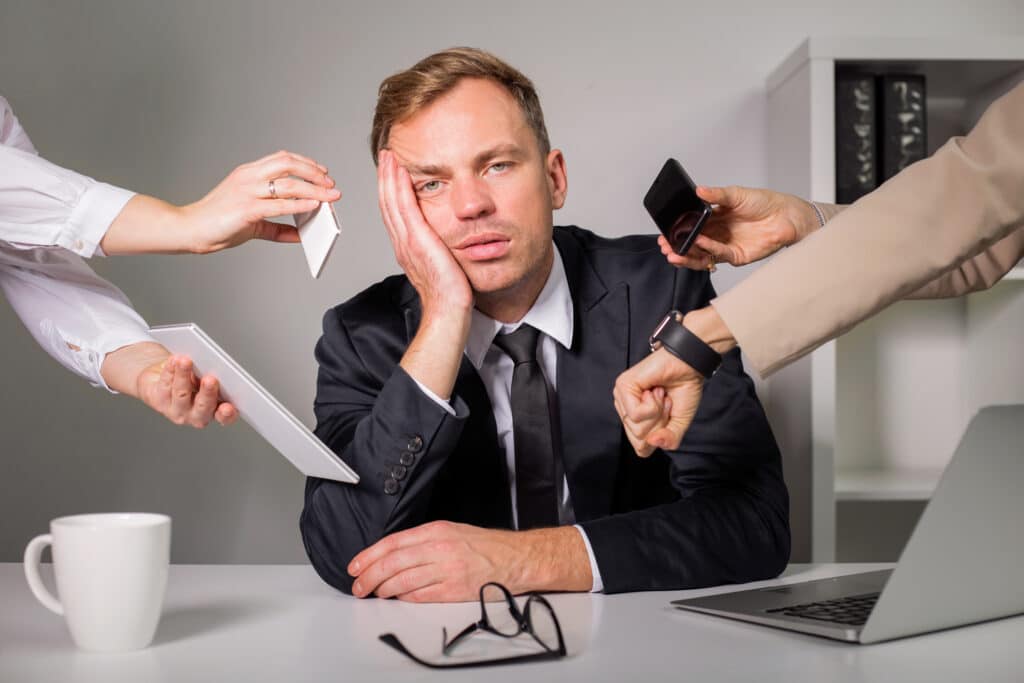 A tired man in a suit sits at his desk, resting his head against his hand, as four coworker hands show tasks that need his attention by holding out an envelope, phone, and watch respectively.
