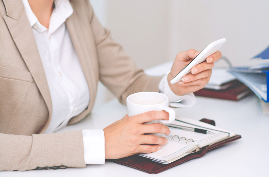 An employee sits at her desk, checking her smartphone while resting a hand on a coffee mug on top of a notebook.