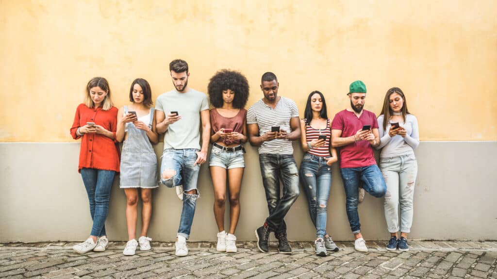 A diverse group of friends stands and rests in a line against a wall. Each of them is looking at a smartphone.