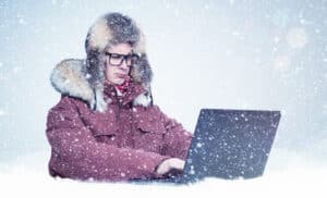 "Email cold leads" concept: a man wearing a parka and wool hat sits in the snow typing on a laptop.