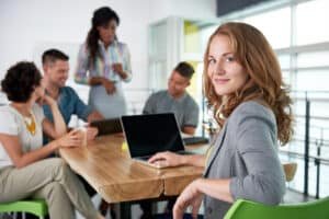 A female Fractional CMO smiles at the camera while sitting at a table with her laptop, a diverse team of marketers across from her.