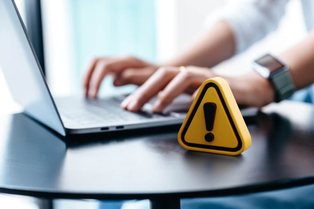 Email safety for digital marketing concept: a yellow exclamation-point warning icon rests on a table beside a marketer using a laptop.