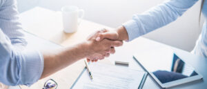 Outsourced marketing concept: two businesspeople shake hands over a desk at the conclusion of a marketing meeting.